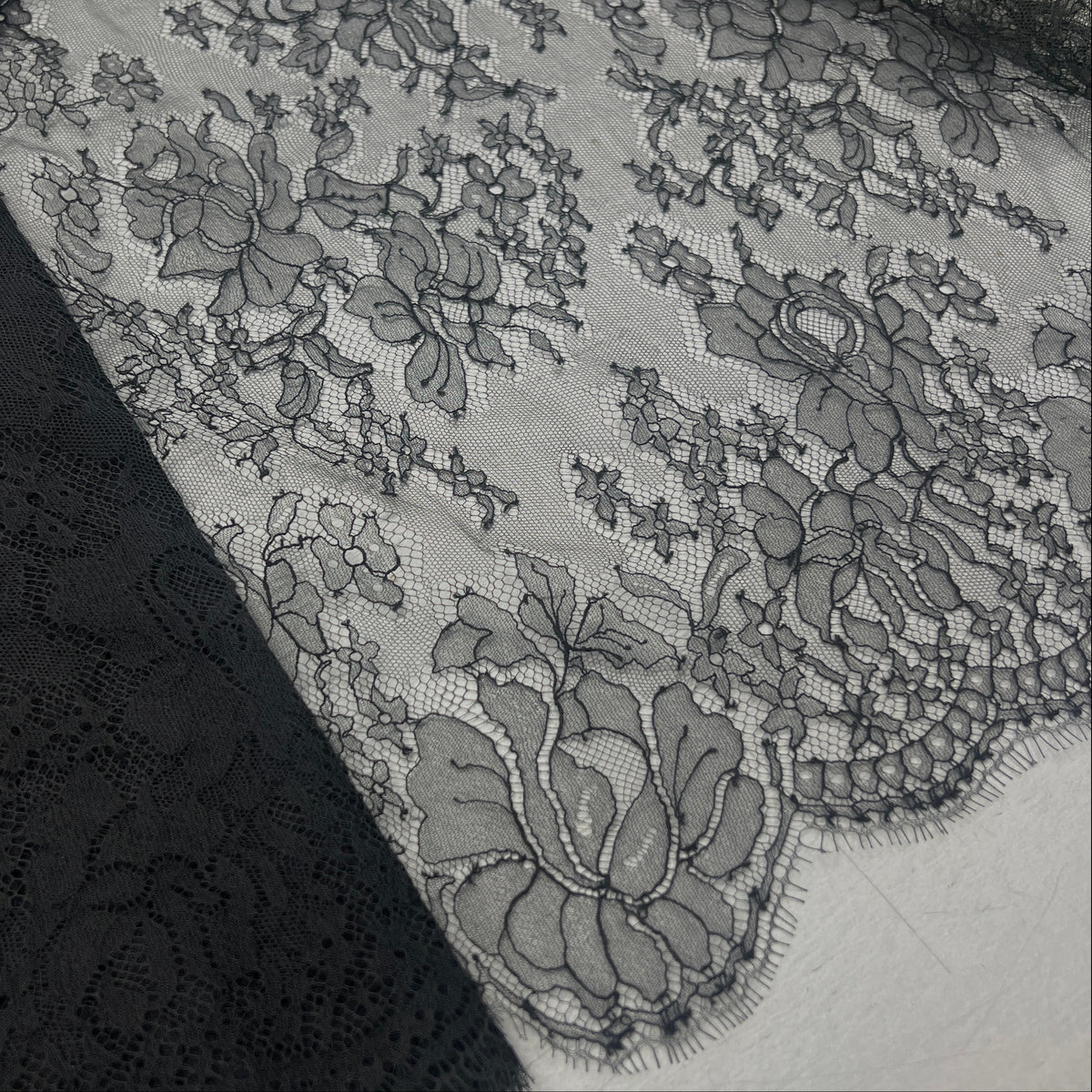 Black floral lace fabric draped over a white surface, showing detailed embroidered lace patterns and sheer mesh.