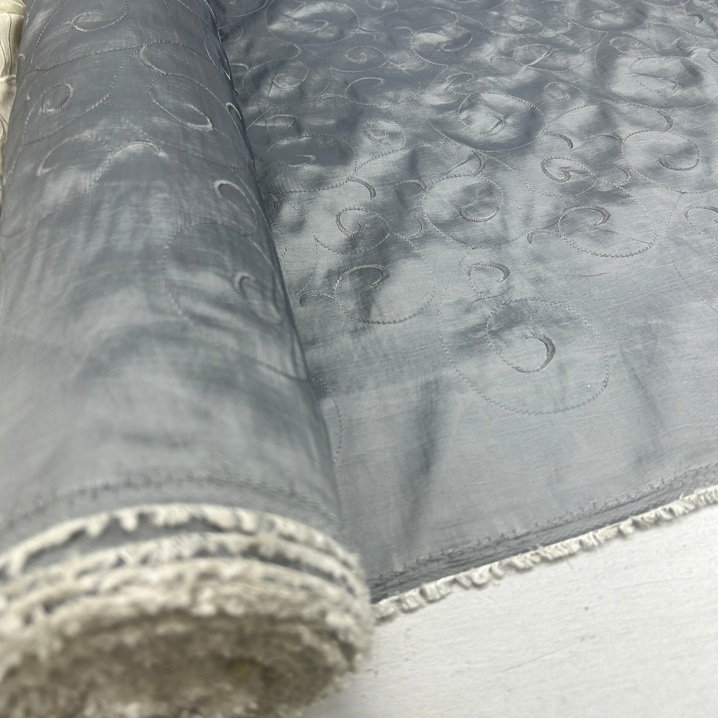 A close-up of a rolled piece of gray fabric with a subtle tonal circular pattern and a frayed white edge, laid on a light surface.