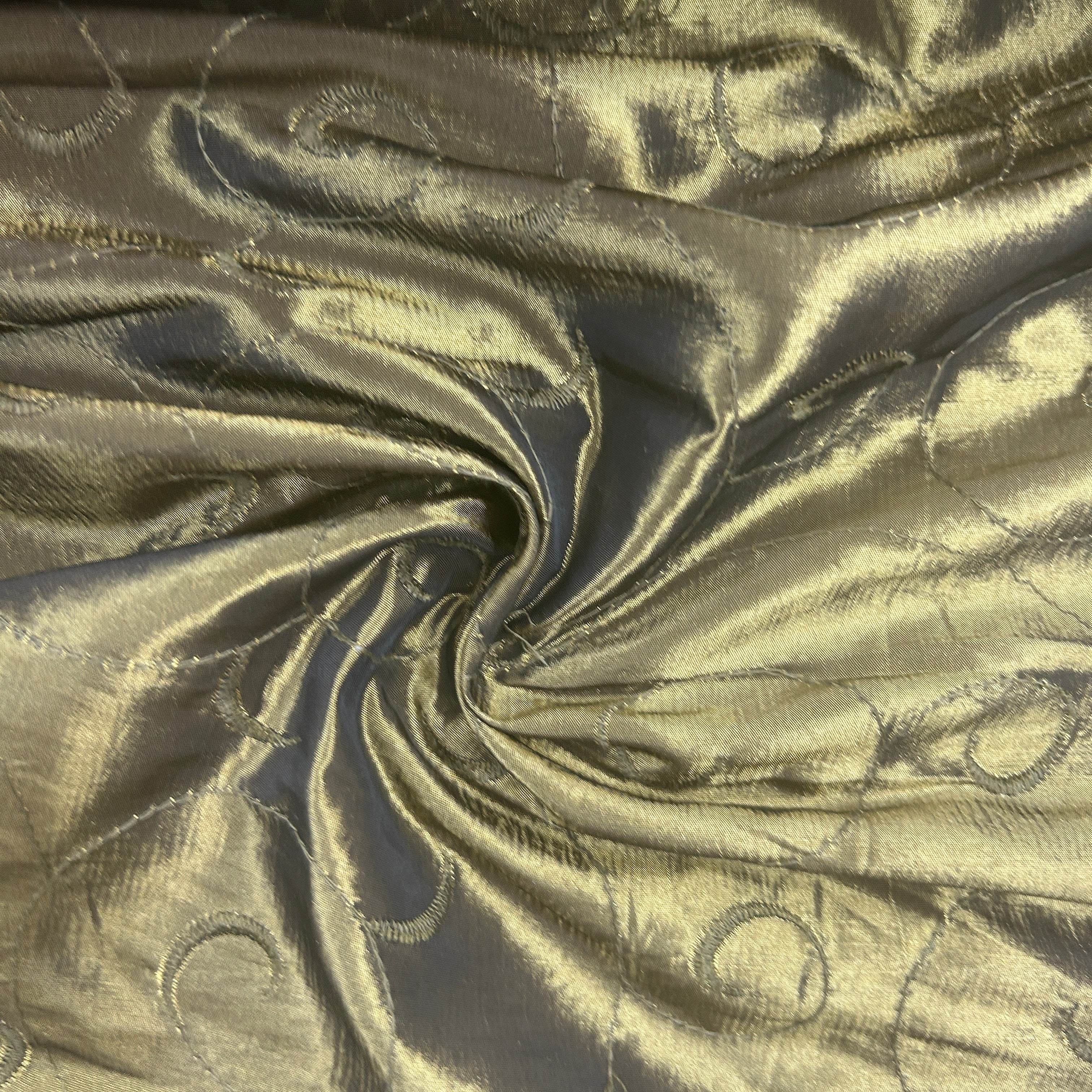 A close-up of shimmering metallic fabric with a warm gold and bronze sheen, gathered in a swirl to show texture and drape. The material has a subtle woven circular pattern.