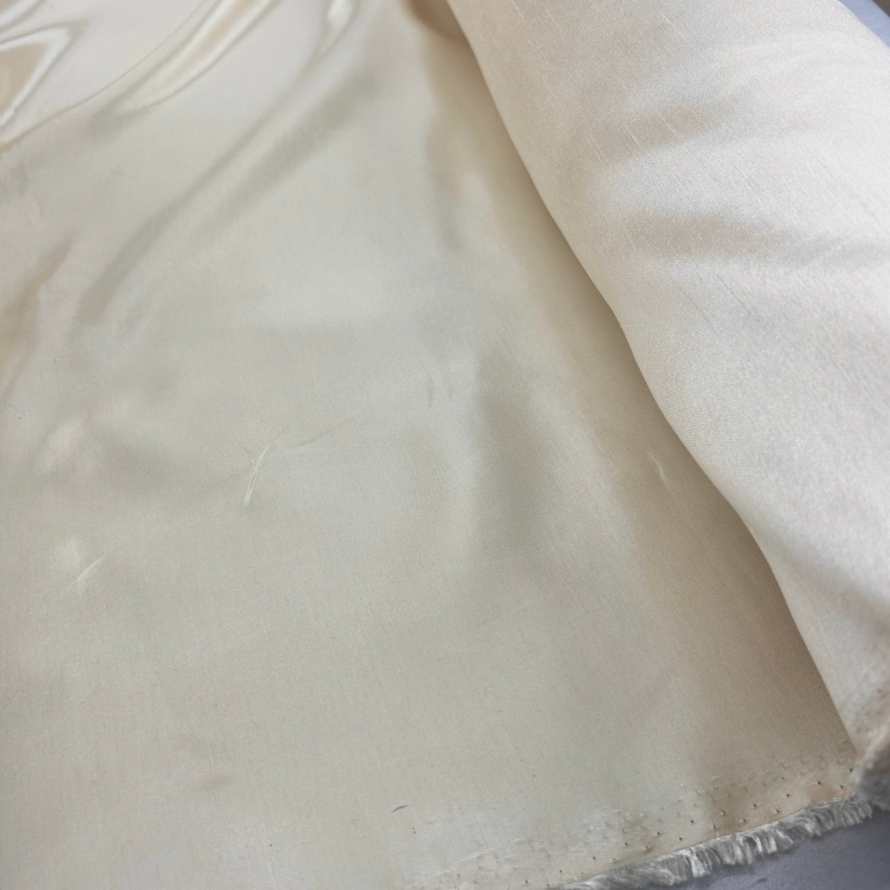 A close-up of a cream-colored fabric, likely silk or satin, draped to show its smooth texture and sheen, with a folded corner revealing the underside.