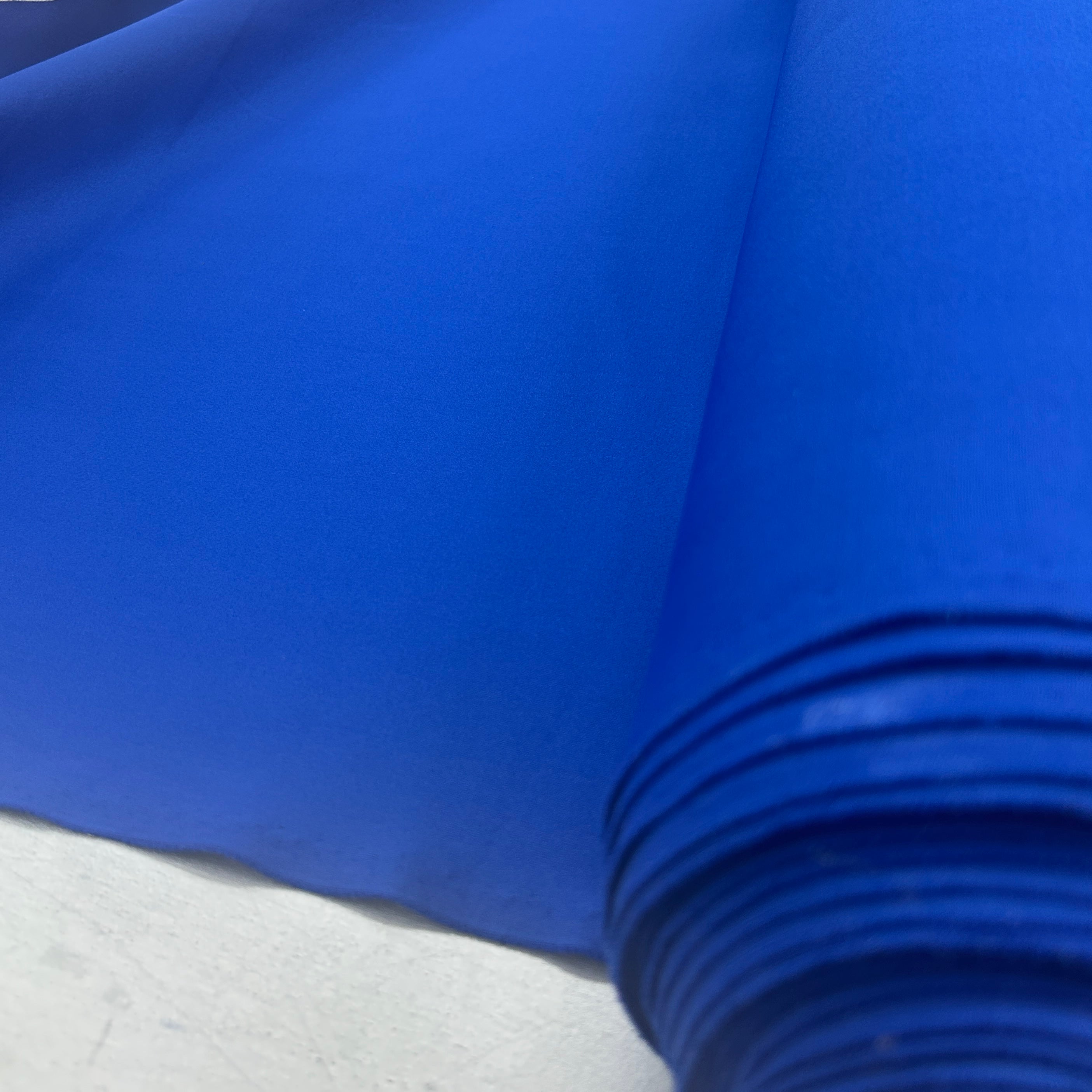 A close-up of a bright royal blue fabric or material roll laid over a light gray surface, with the fabric draped across the frame and stacked layers visible at the bottom right.