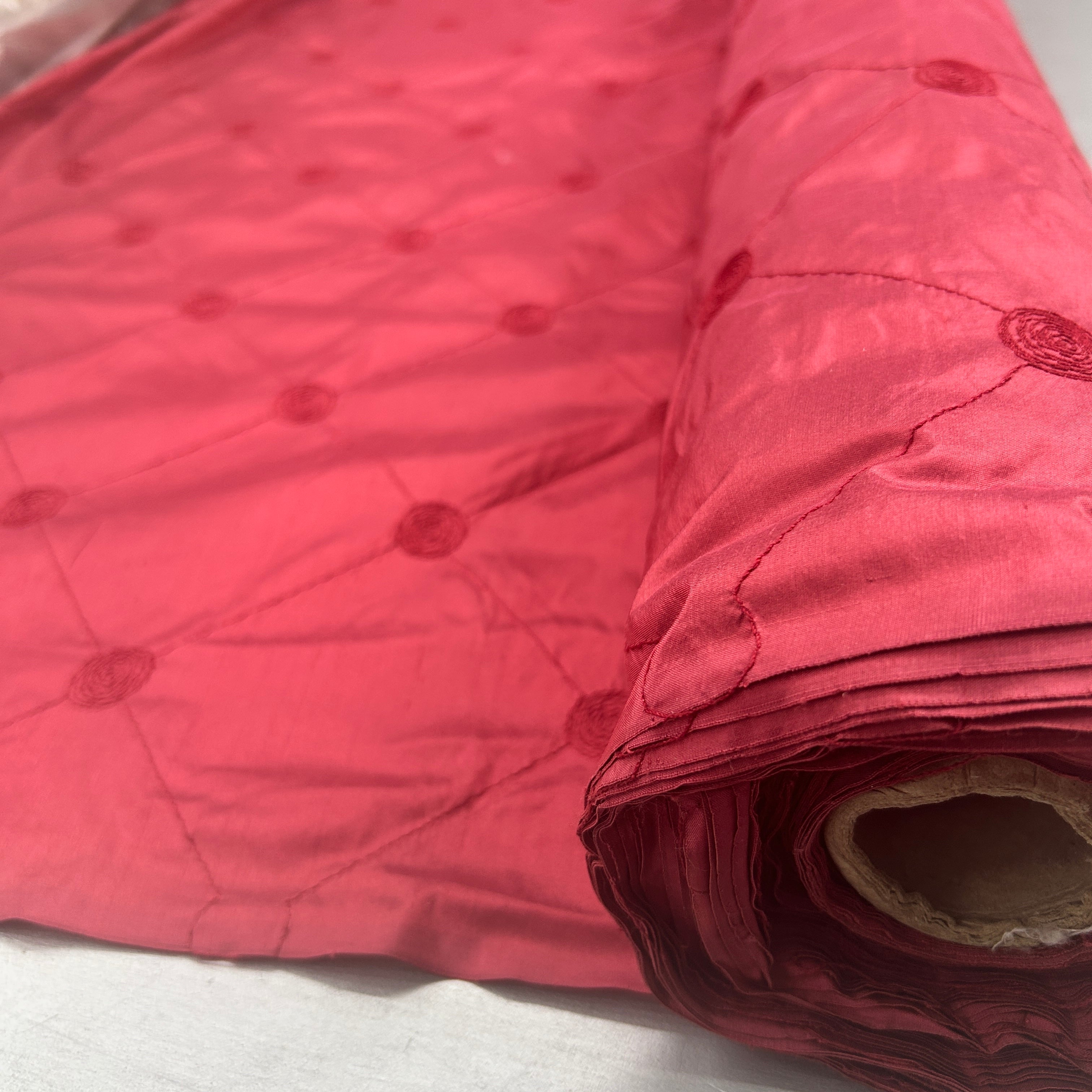 A close-up of a rolled piece of shiny pink fabric with a quilted diamond pattern and tonal circular motifs, laid on a light surface.