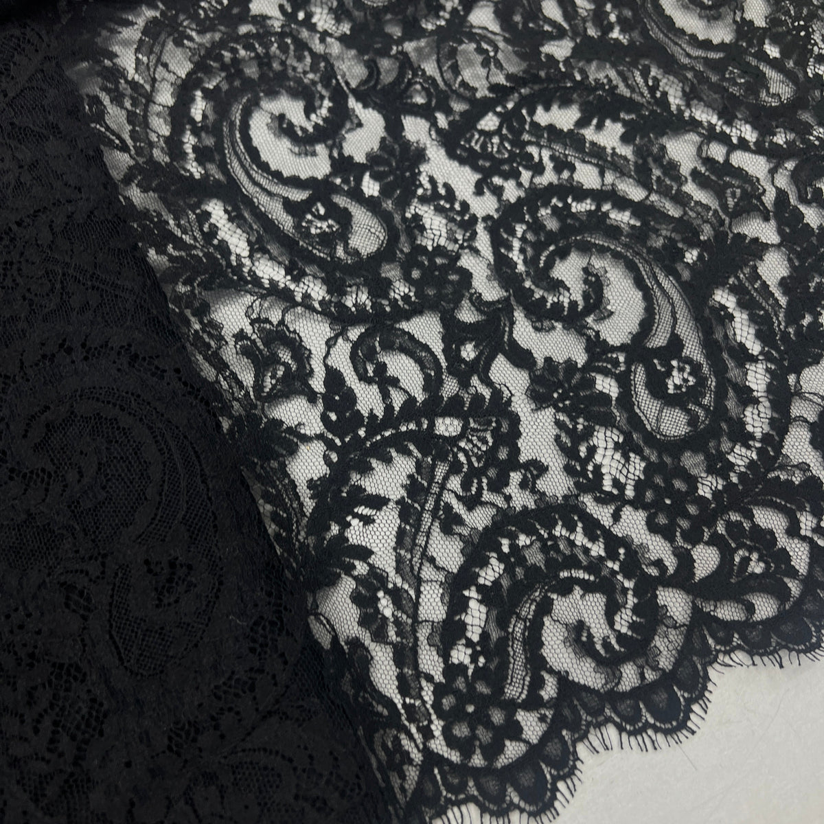 Black lace fabric with an intricate floral and scroll pattern, shown draped over a white surface.