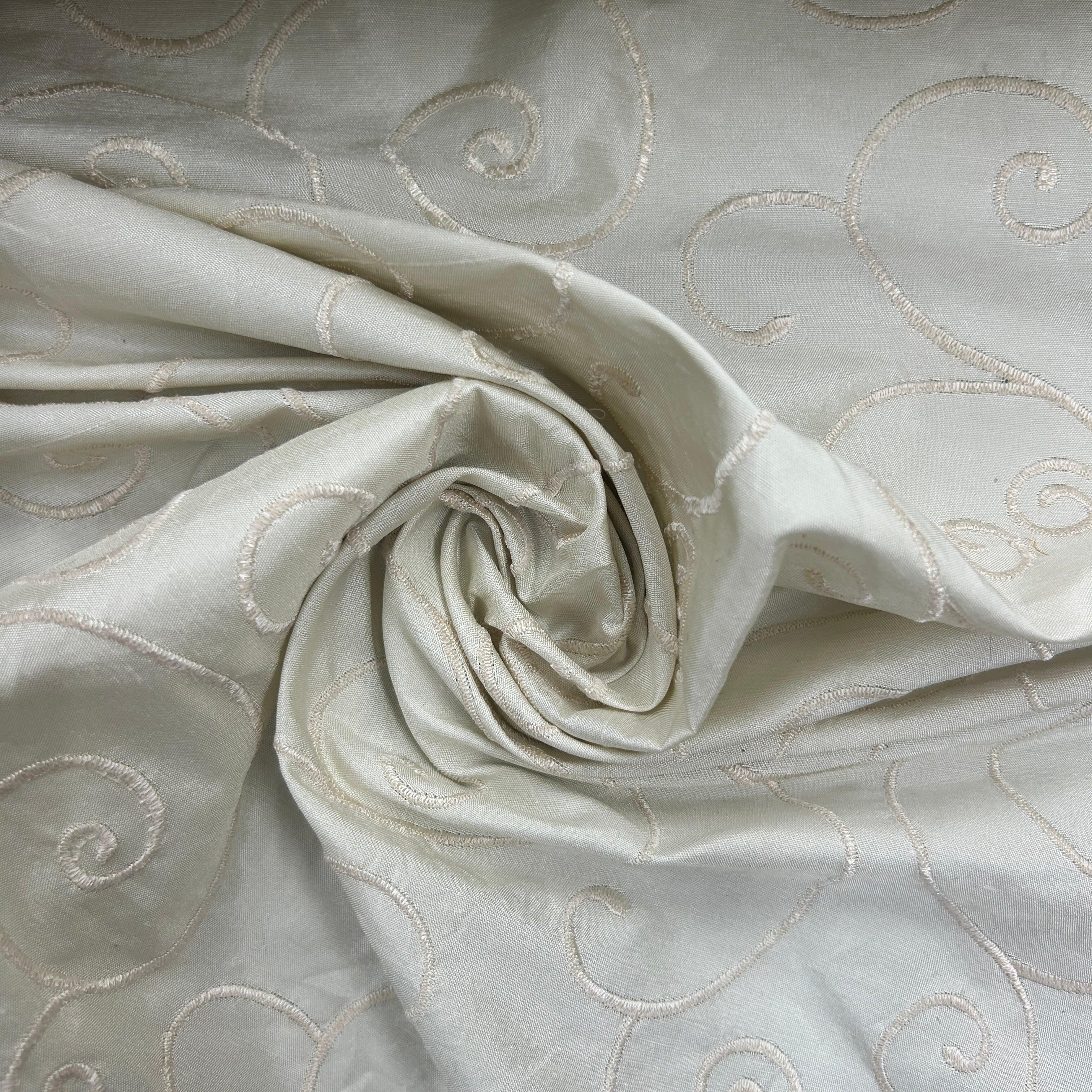 A close-up of a light ivory or off-white fabric with a subtle sheen, featuring a swirling, draped texture and delicate metallic gold embroidered swirl patterns.