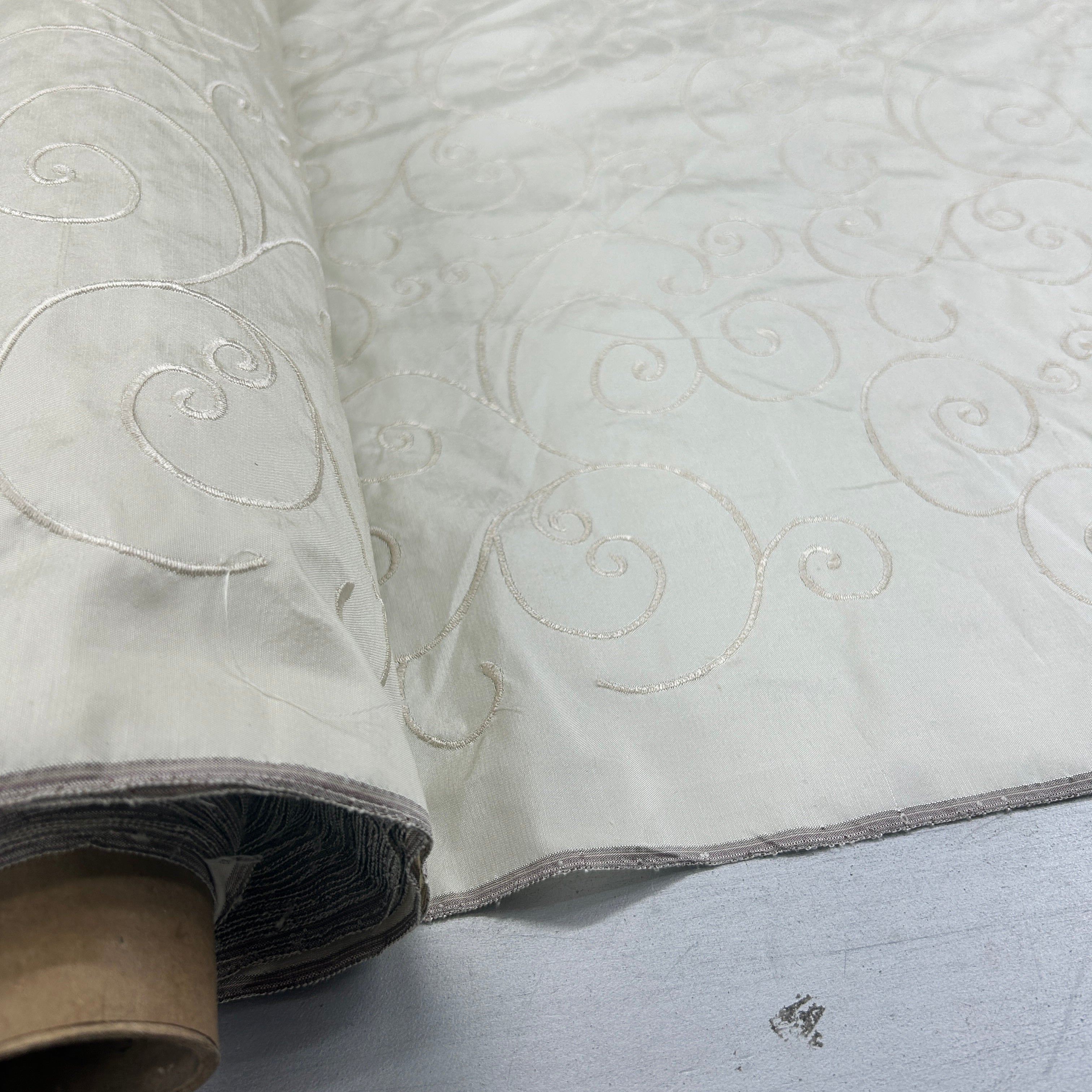 A close-up of a rolled fabric or wallpaper sample in warm off-white/ivory, featuring a repeating raised swirl pattern and a slightly textured surface, shown on a light gray surface.