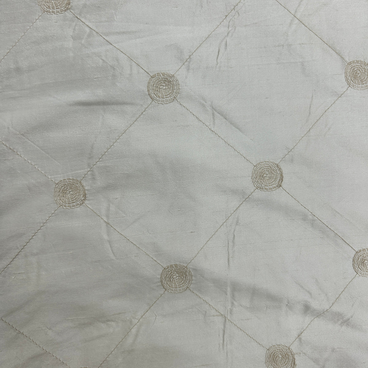 A close-up of a light gray or off-white fabric with a subtle woven texture, featuring a stitched diamond grid pattern and circular medallion motifs at the intersections.