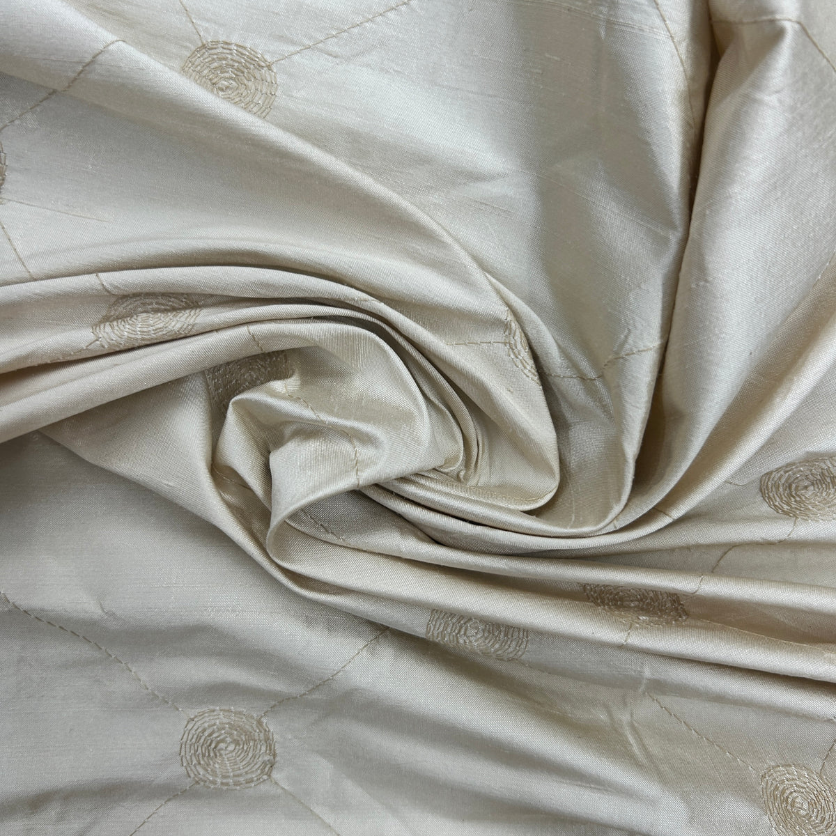 A close-up of draped cream-colored fabric with a subtle sheen and small circular woven motifs, arranged in soft folds.