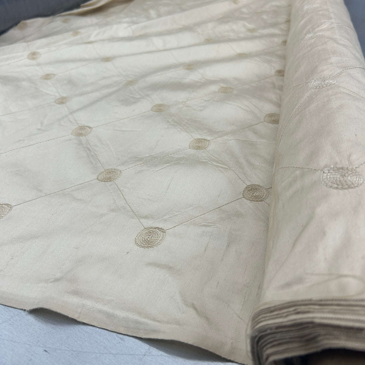 A close-up of a cream-colored fabric, likely silk or satin, featuring a subtle sheen and a repeating pattern of small circular motifs connected by thin lines. The fabric is draped to show its texture and is rolled on the right side.