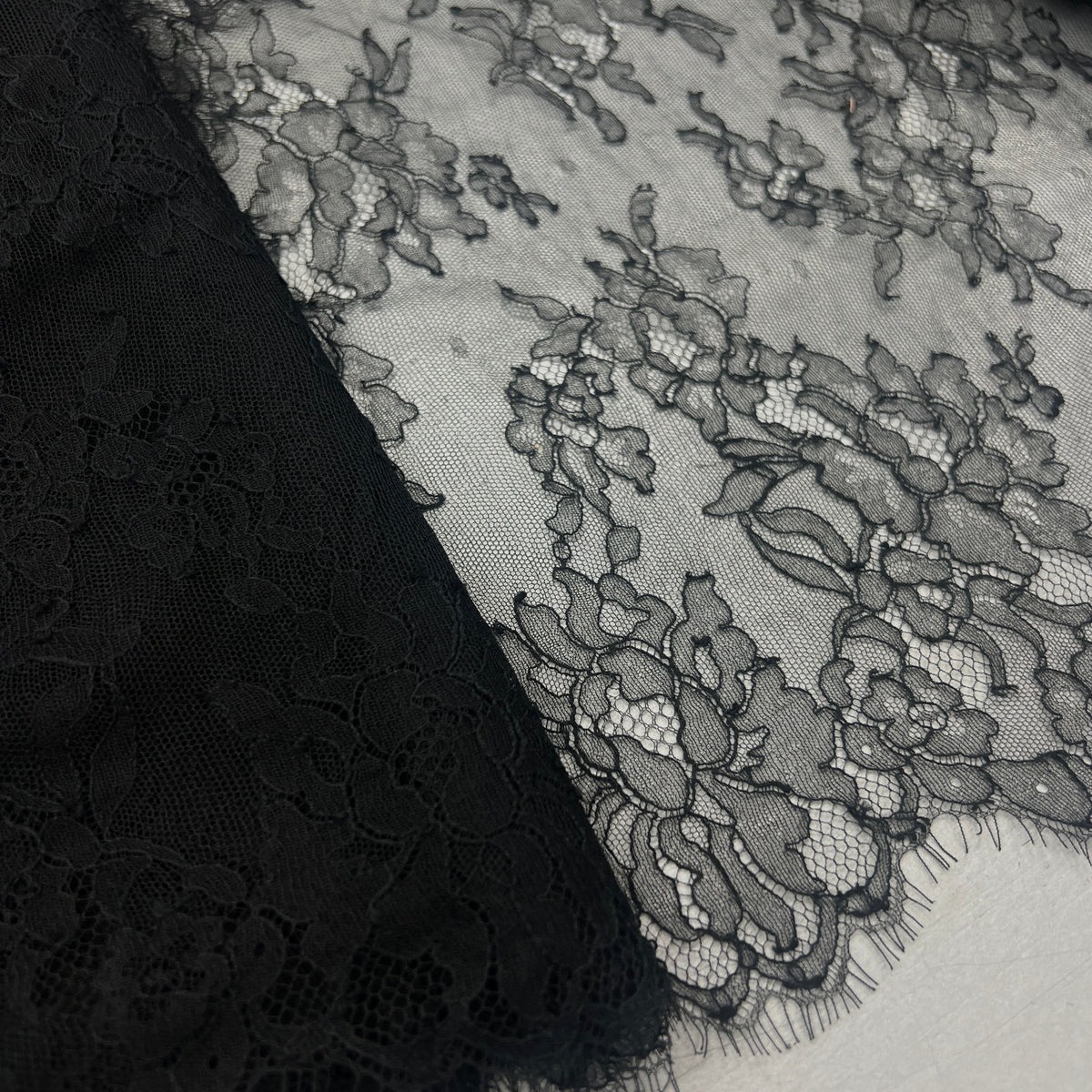 Black floral lace fabric with delicate mesh and embroidered details, shown draped over a white surface.