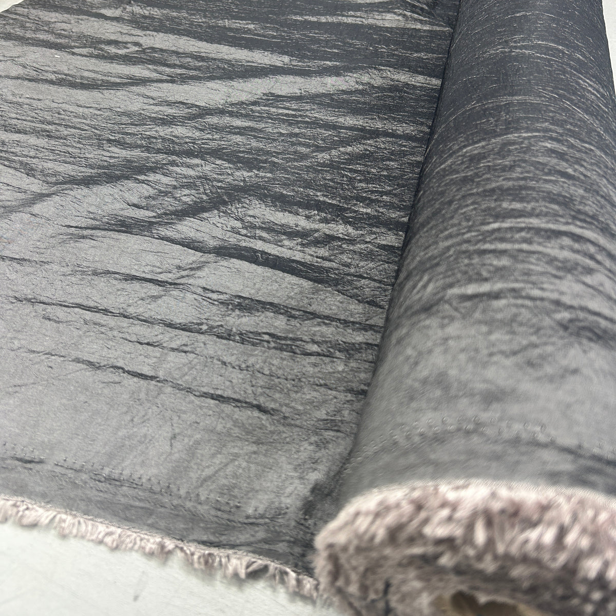 A close-up of a dark gray textured fabric with a crinkled, metallic-like surface and a fringed edge, shown partially rolled on a white surface.