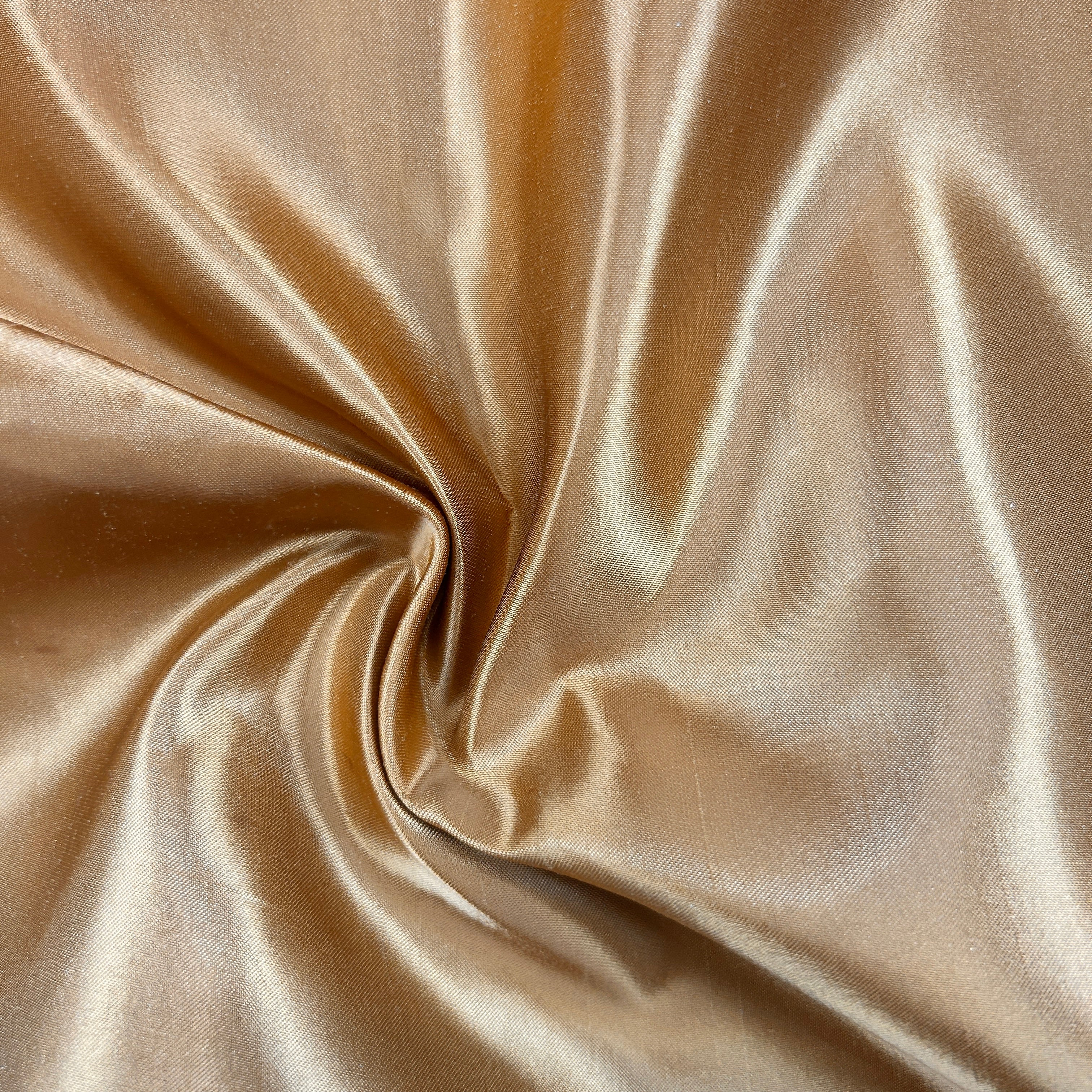 A close-up of draped satin fabric in a warm champagne-gold color, arranged in a central swirl to show the material's sheen and texture.