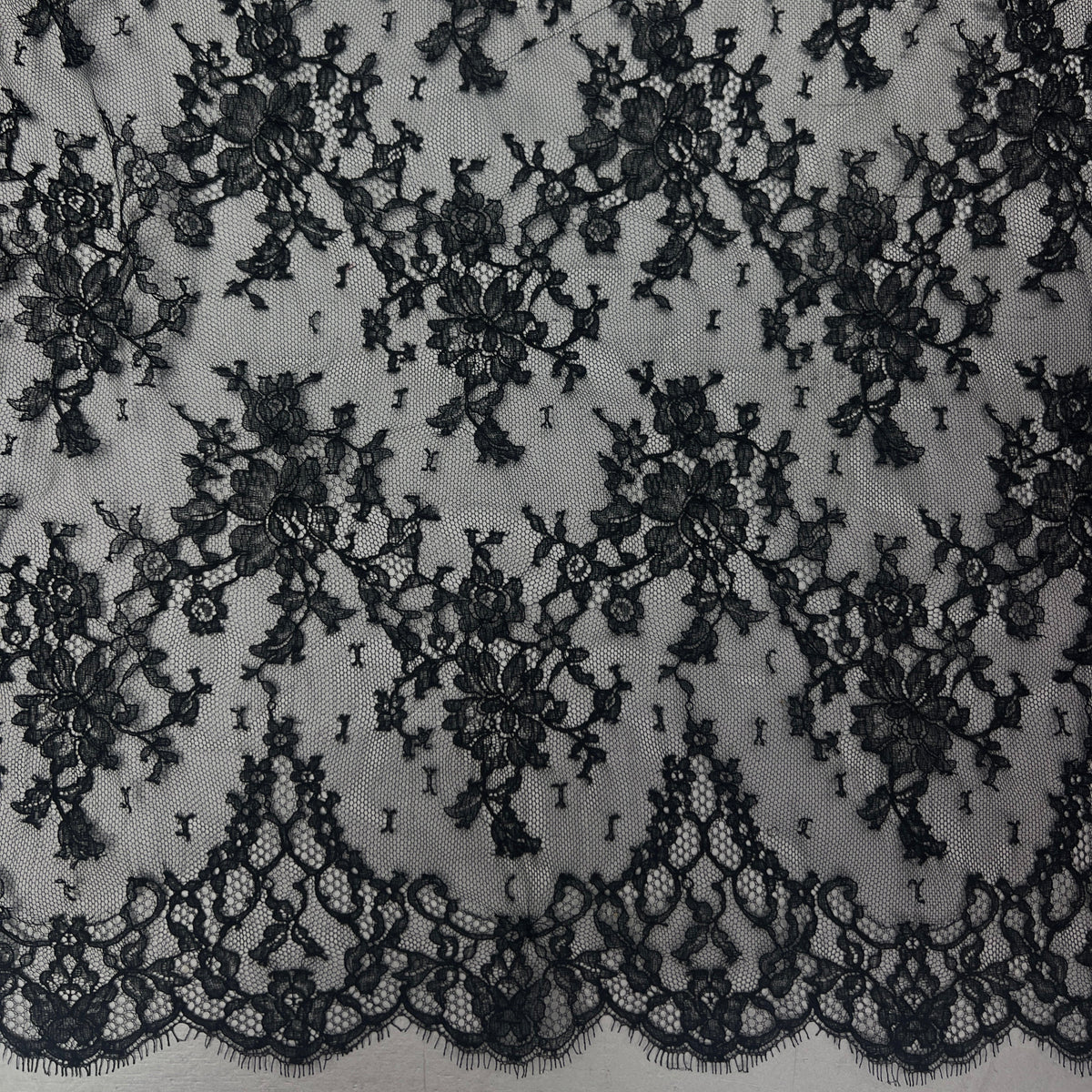 Black floral lace fabric with an intricate embroidered pattern and a scalloped lace border, shown laid flat against a light gray background.