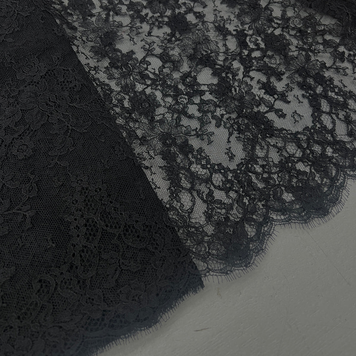 Black lace fabric with a floral pattern and scalloped edge, laid flat on a light gray surface.