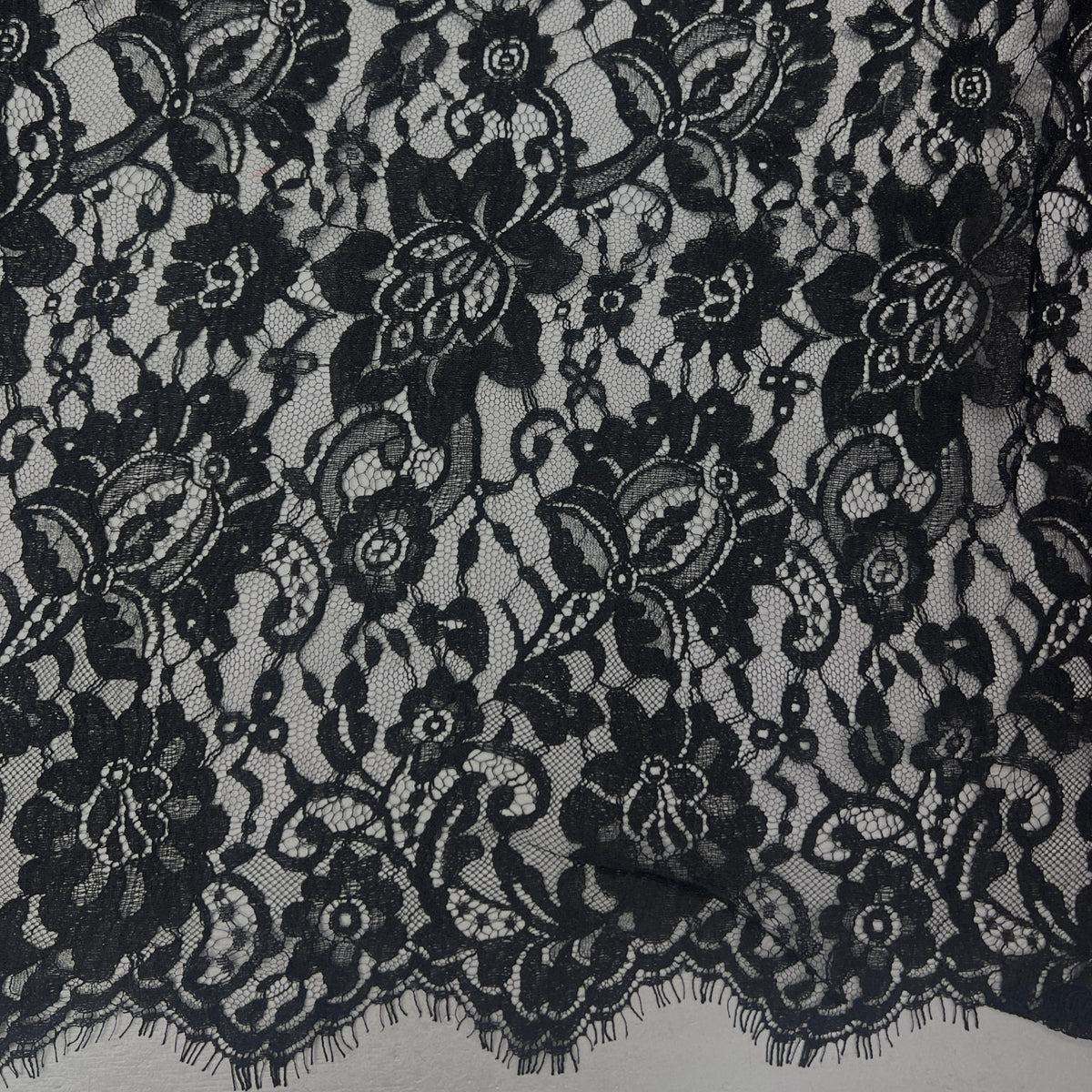 Black floral lace fabric with intricate embroidered details and a scalloped edge, shown in a close-up texture view.