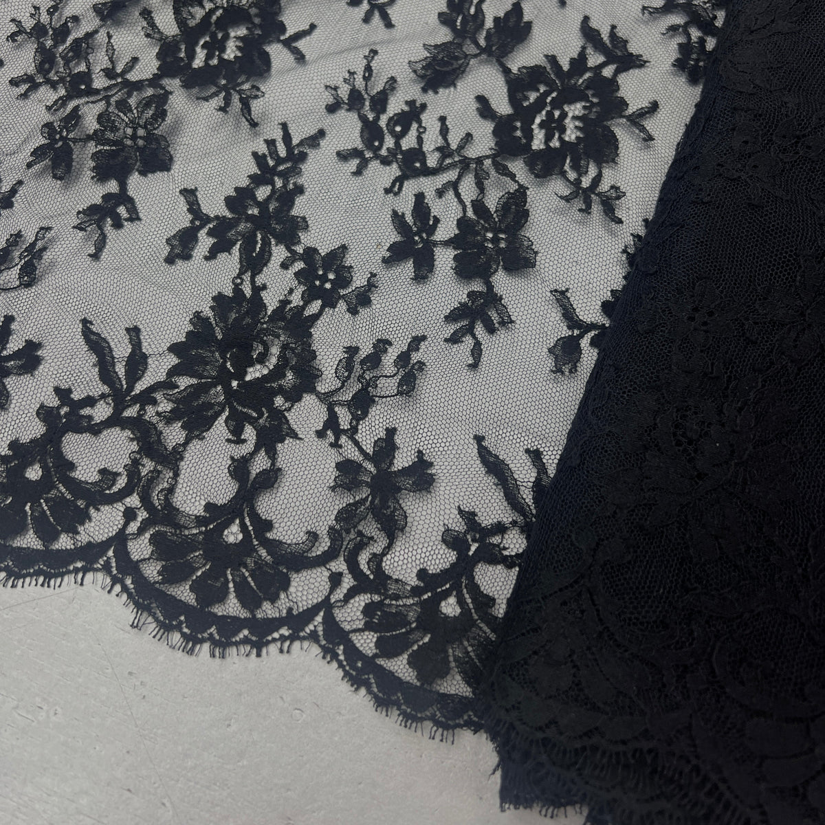 Black floral lace fabric with embroidered floral motifs and scalloped edges, shown draped over a light neutral surface.