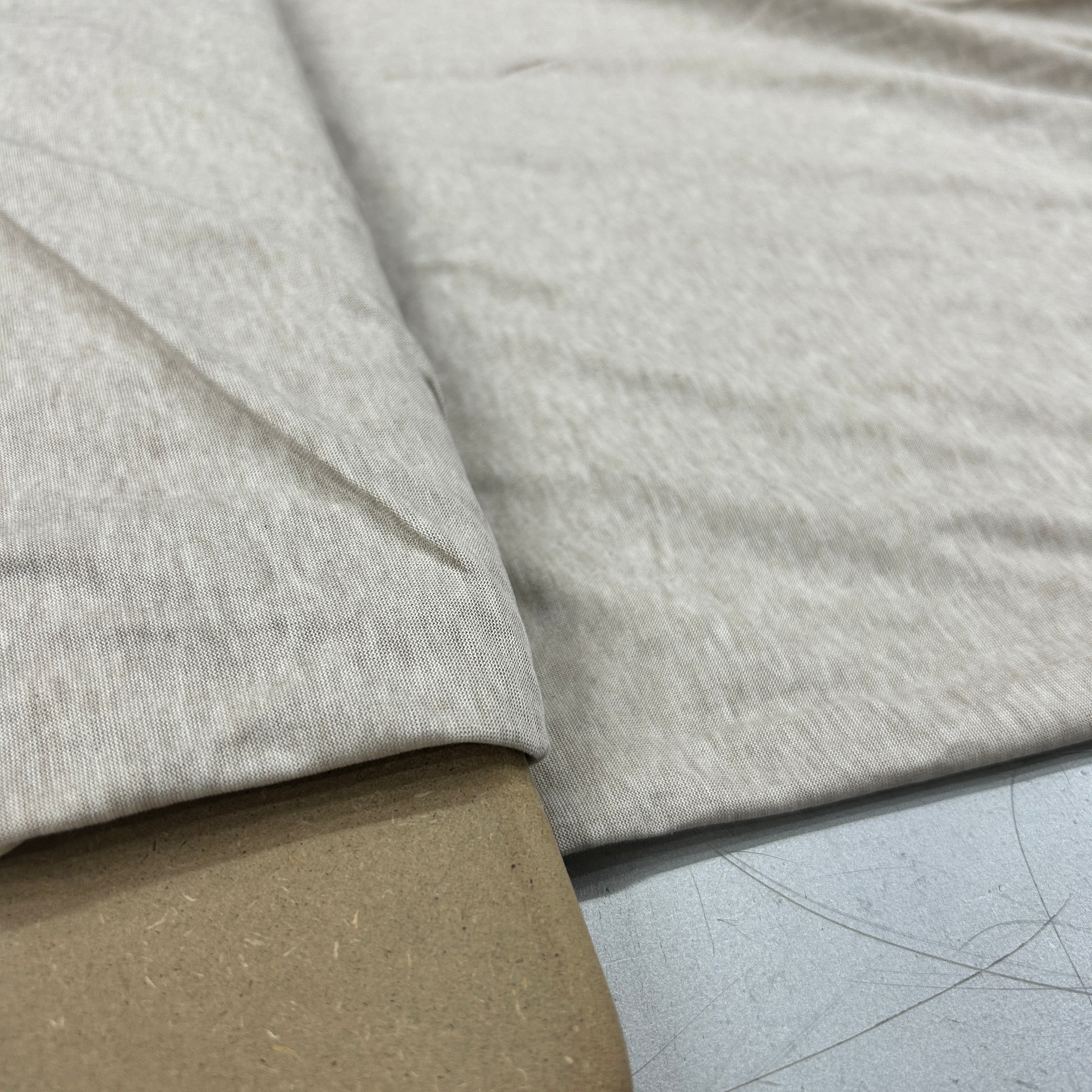 A close-up of light heather gray knit fabric draped over a brown cardboard surface, showing the texture and color of the material.