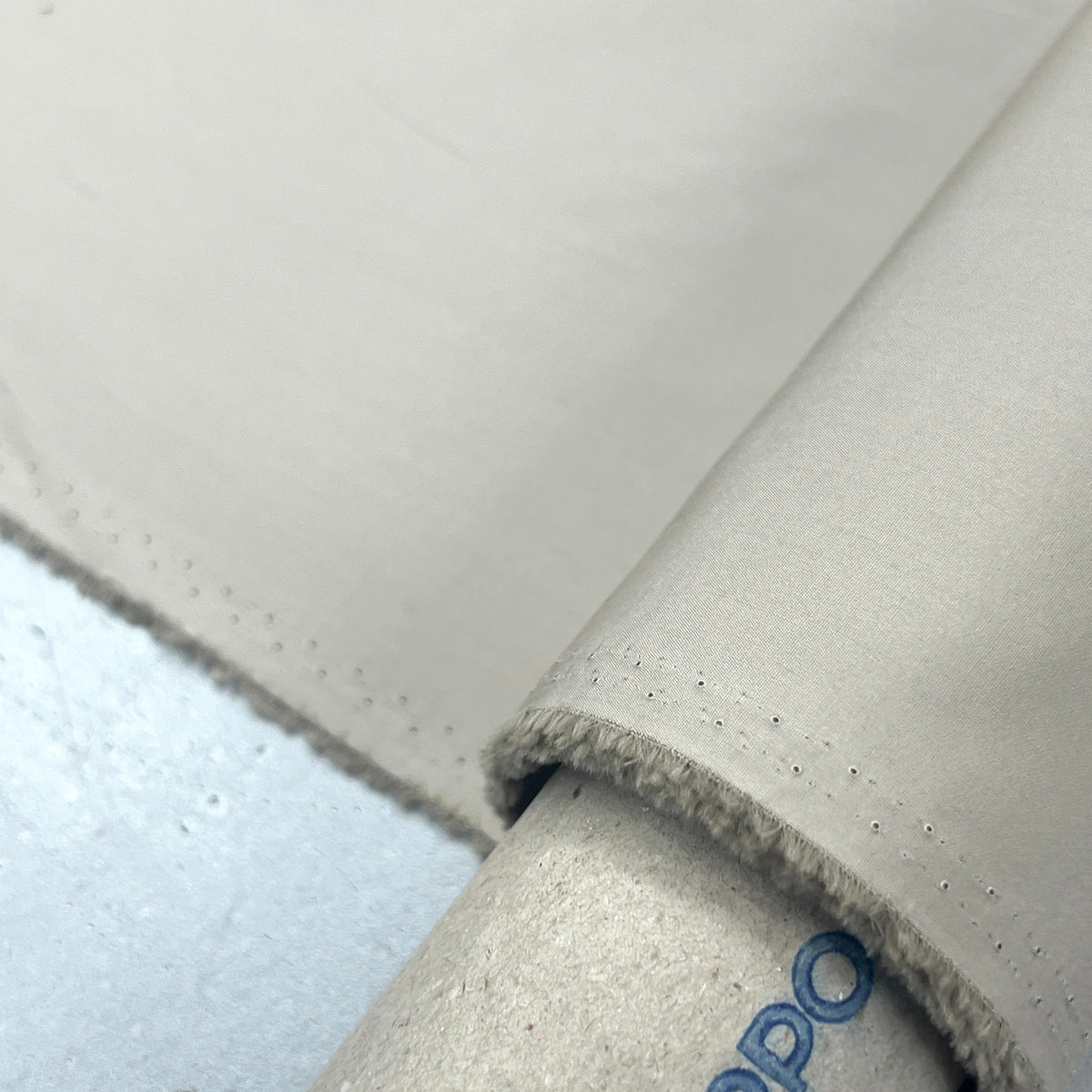 A close-up of a light beige fabric roll with a matching flat sheet laid out, showing a rolled edge with dense short nap and a visible blue printed brand name 'RUPPO' on the tube.