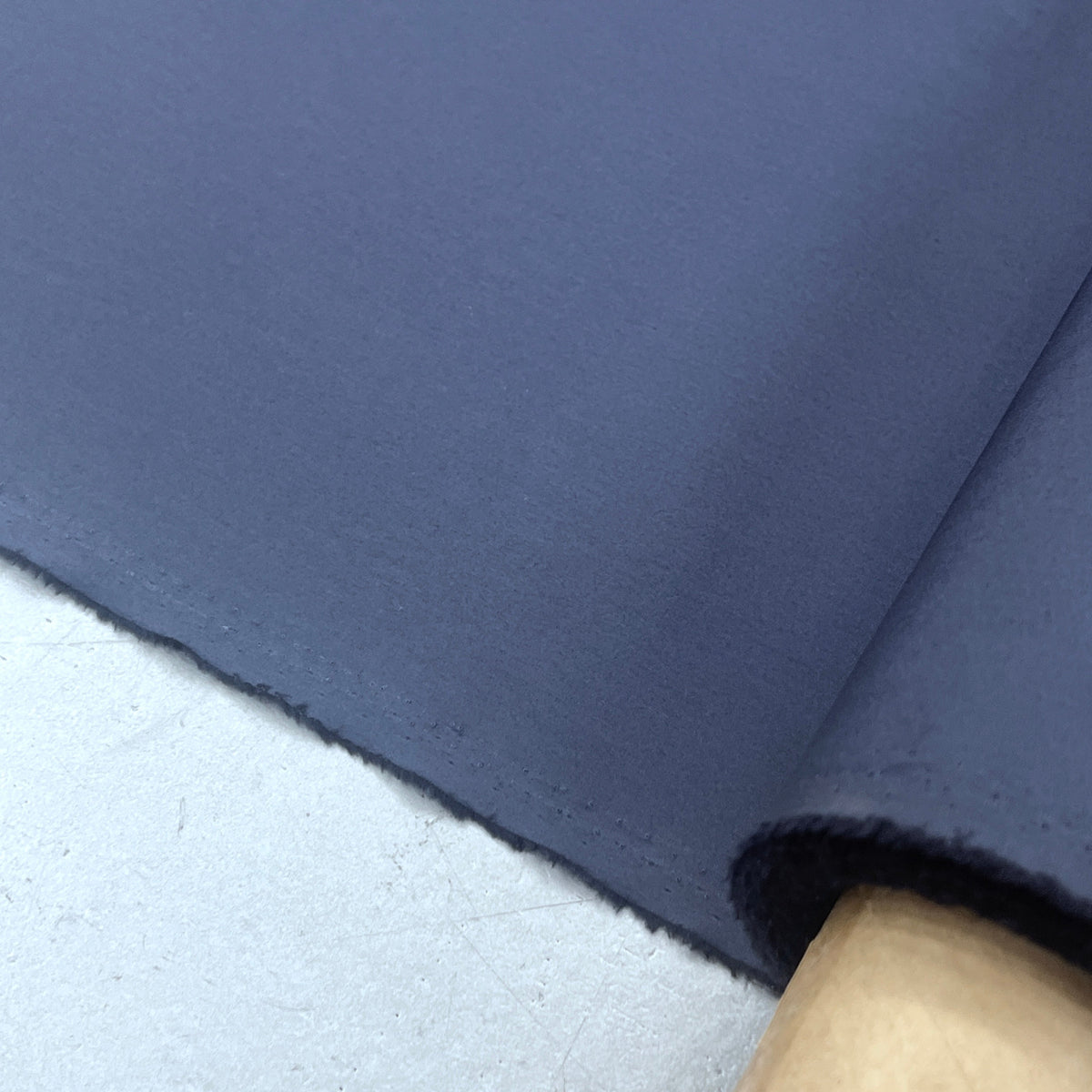 A close-up of a dark slate blue fabric roll, showing its texture and color, resting on a light gray surface.