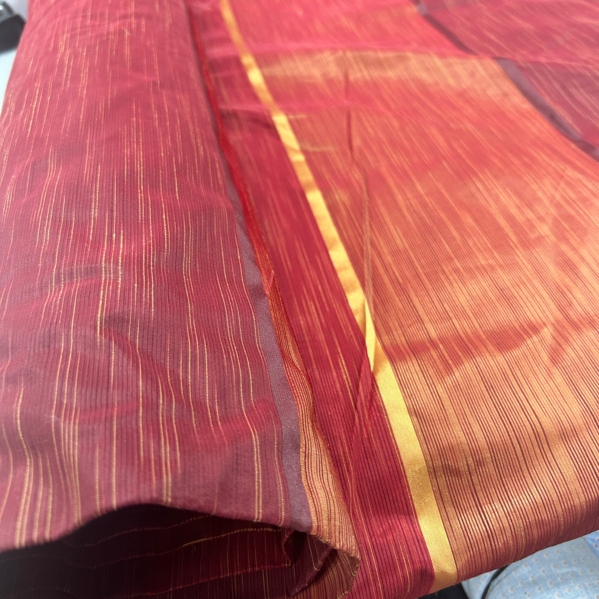 A close-up of a folded and draped red and orange silk saree with fine gold zari stripes.