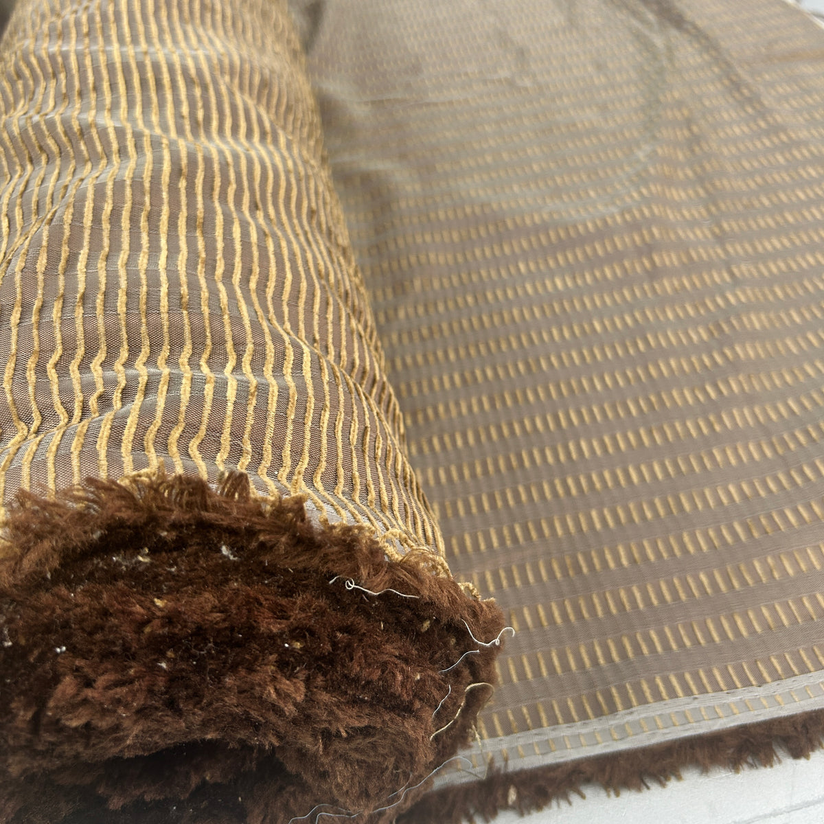 A close-up of a folded rug showing its striped pattern in brown and beige, with a plush dark brown shaggy pile on one side.
