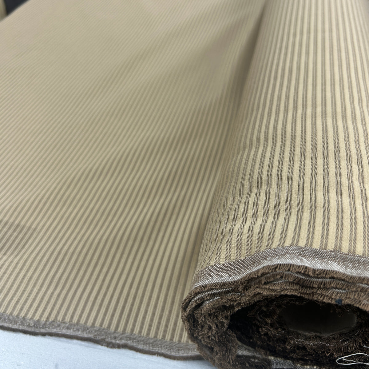 A close-up of a rolled piece of fabric with fine vertical stripes in beige and brown, showing the textured woven edge.