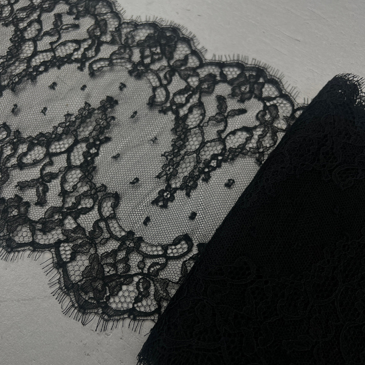 Black lace fabric with floral patterns and a solid black textured trim, displayed on a light gray background.