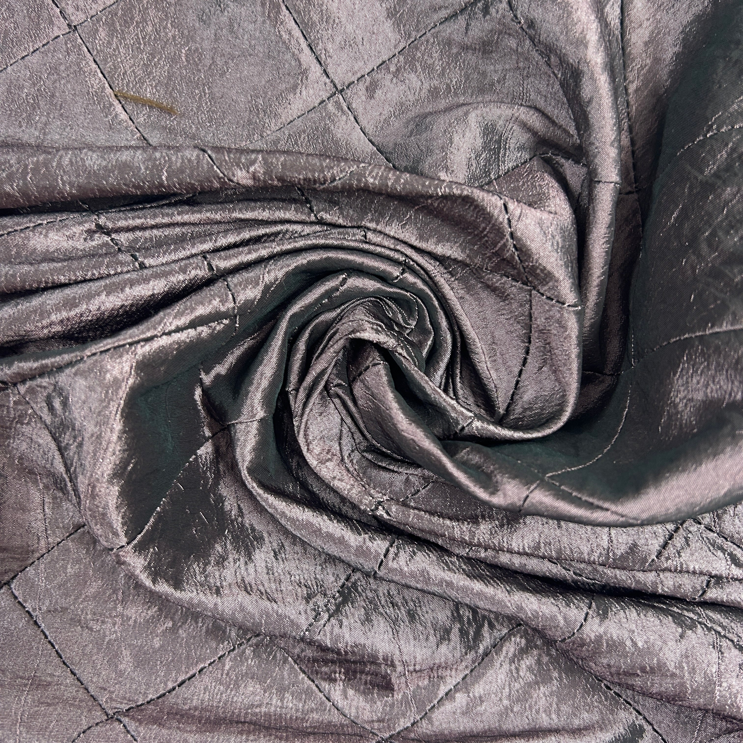 A close-up of draped metallic fabric with a quilted diamond pattern, shown in a spiral fold to highlight its texture and sheen.