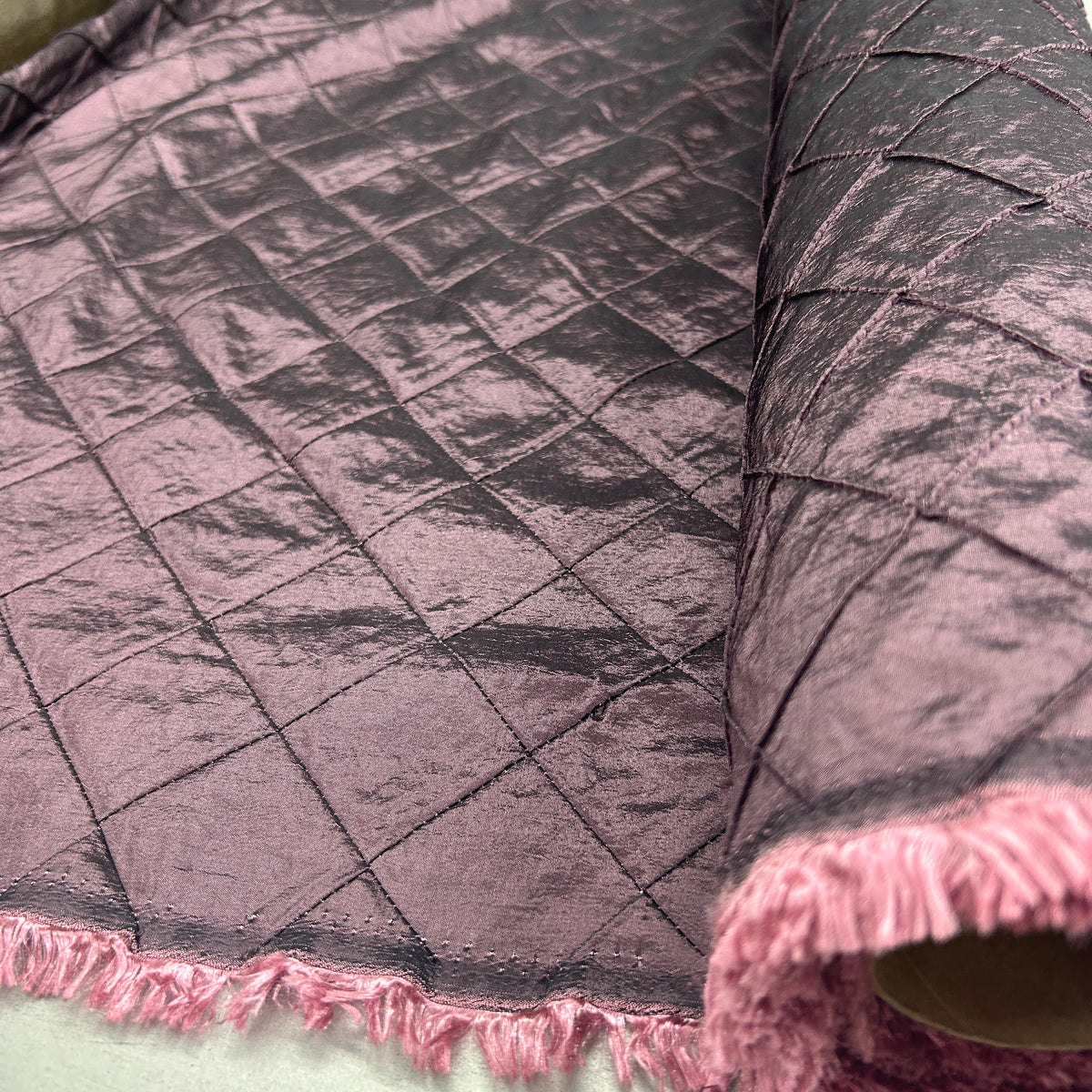 A close-up of a dark charcoal gray quilted velvet fabric with pink fringe trim along the edge, shown rolled out and folded.