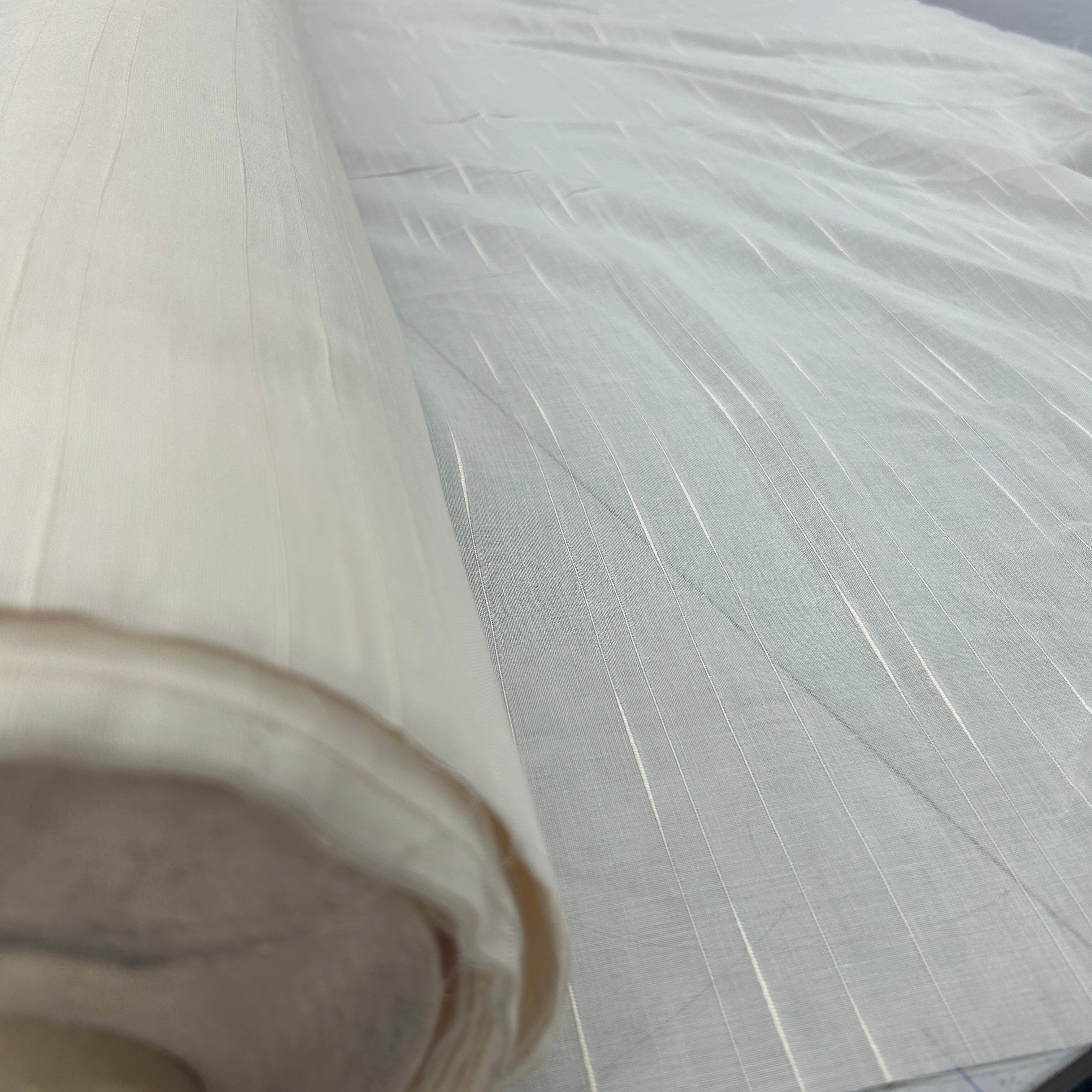 A close-up of a rolled off-white fabric or textile with a subtle woven texture and fine vertical striations, shown against a matching light background.
