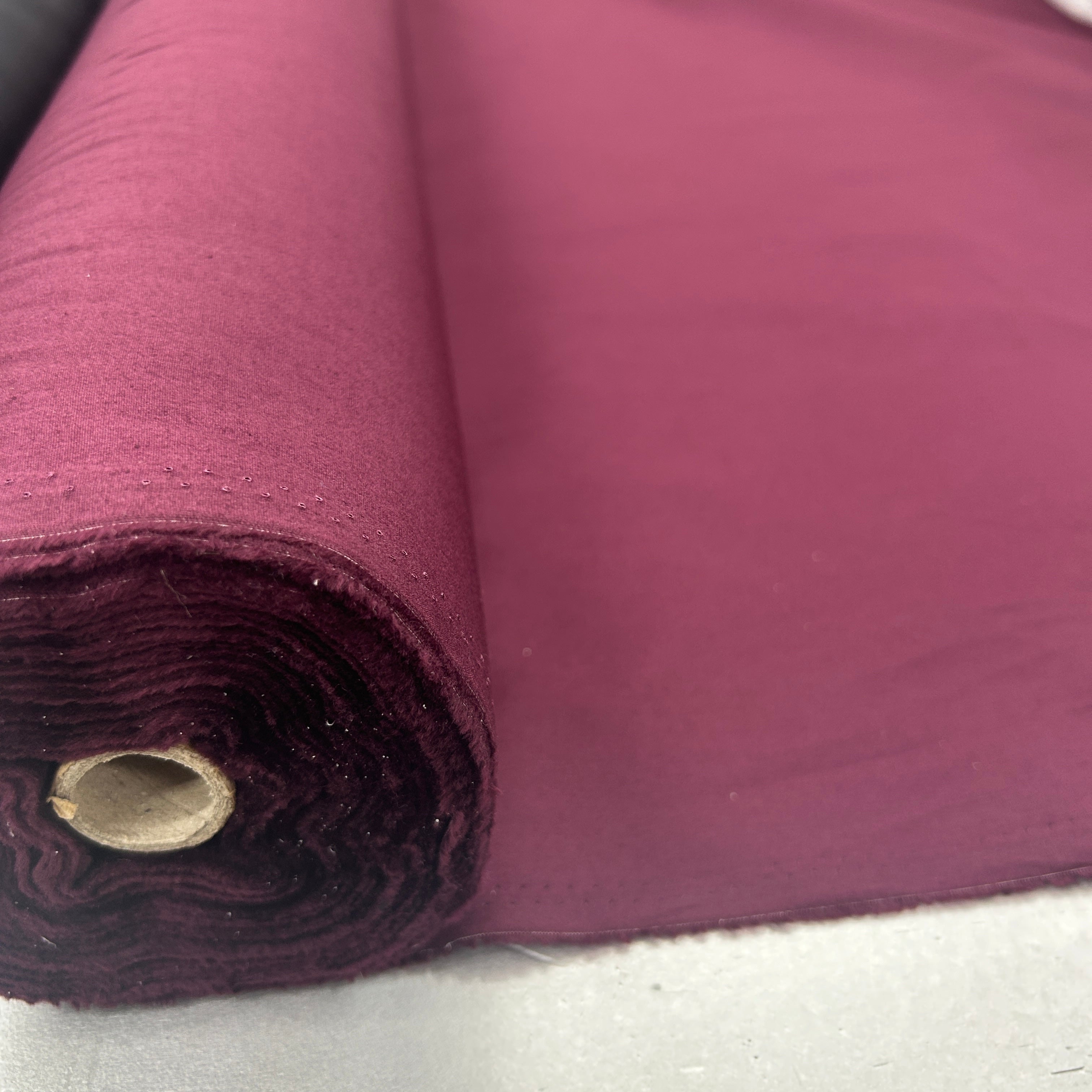 A close-up of a roll of deep magenta fabric, partially unrolled to show its texture and color, set against a neutral light gray background.