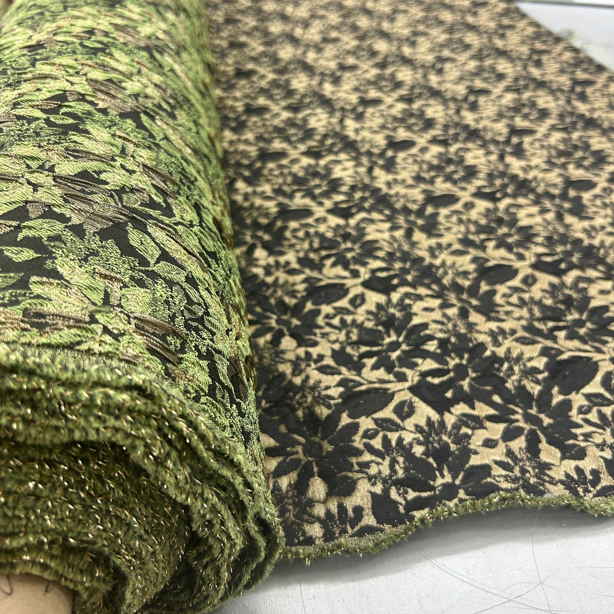 Roll of green and black floral brocade upholstery fabric with intricate woven leaf patterns.