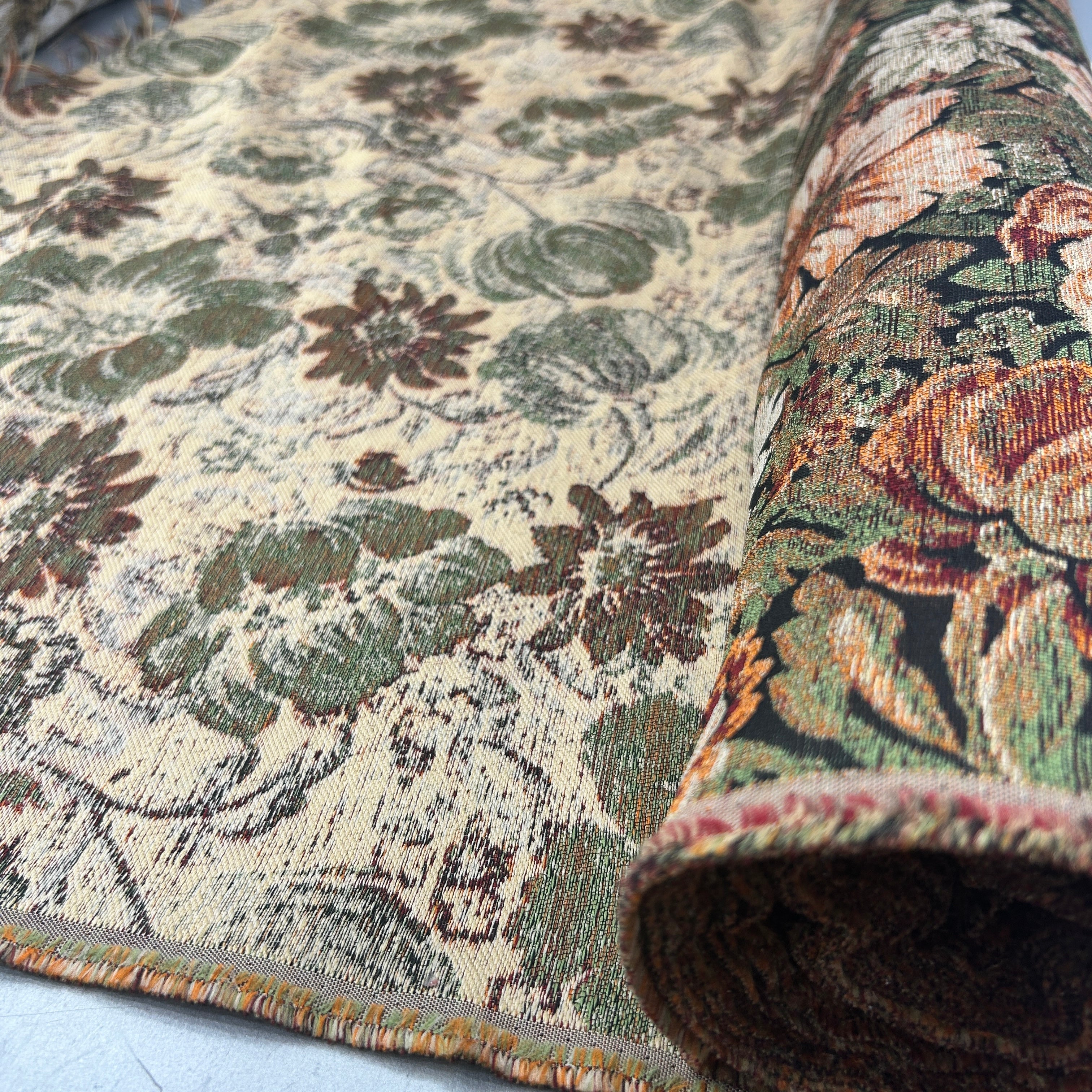 A close-up of a patterned area rug with one end rolled back to show the underside and pile. The rug features a vintage floral design in muted tones of green, brown, and rust orange on a warm beige background.