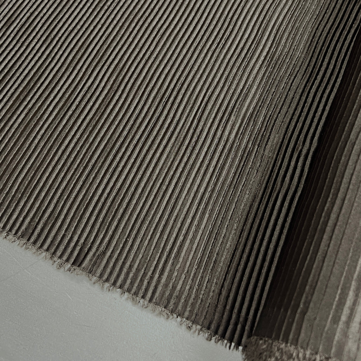 A close-up of a dark gray pleated fabric or material sample with fine fringe along the edge, shown against a light neutral background.