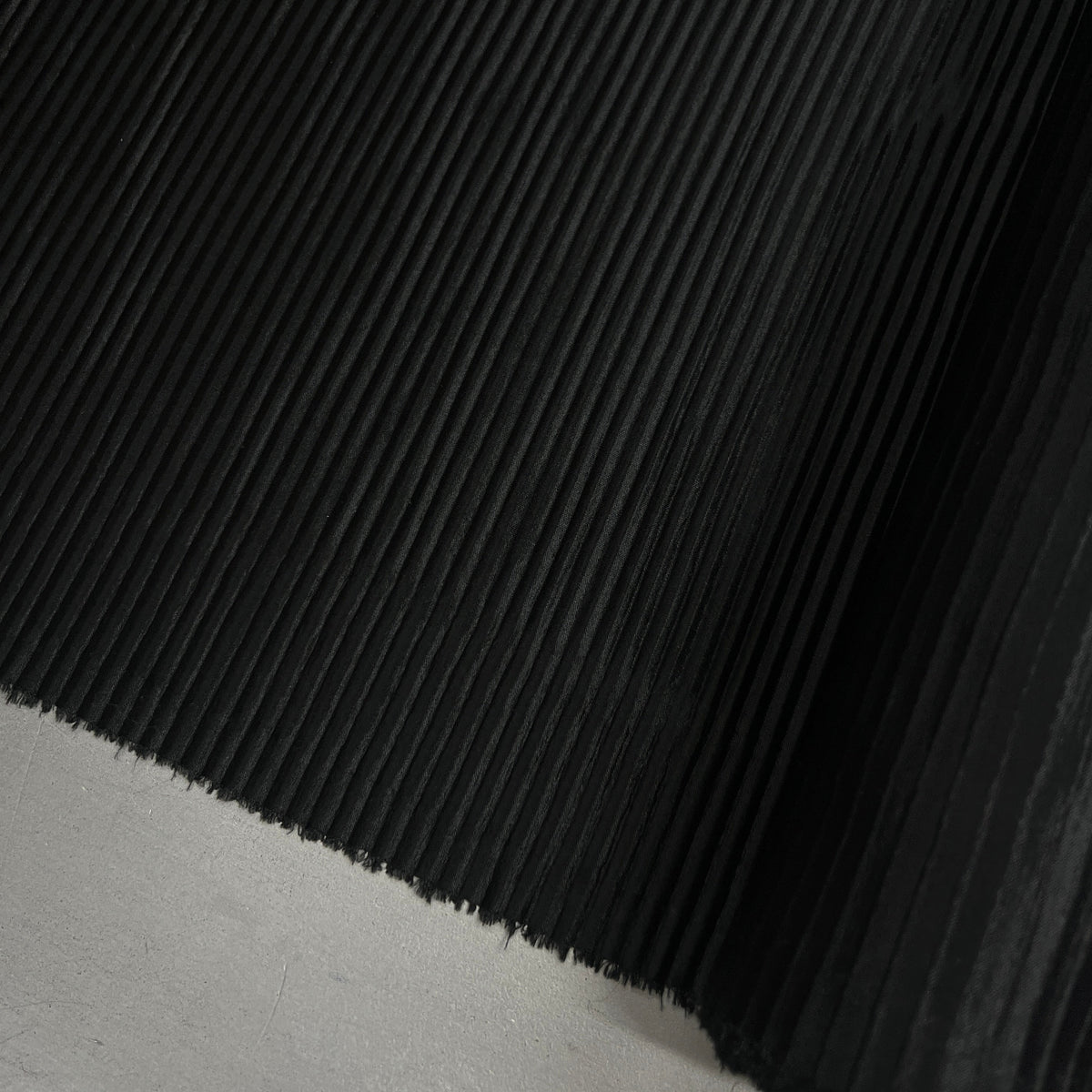 A close-up of a black pleated fabric, showing the fine pleated texture and folded edges, resting on a light gray surface.