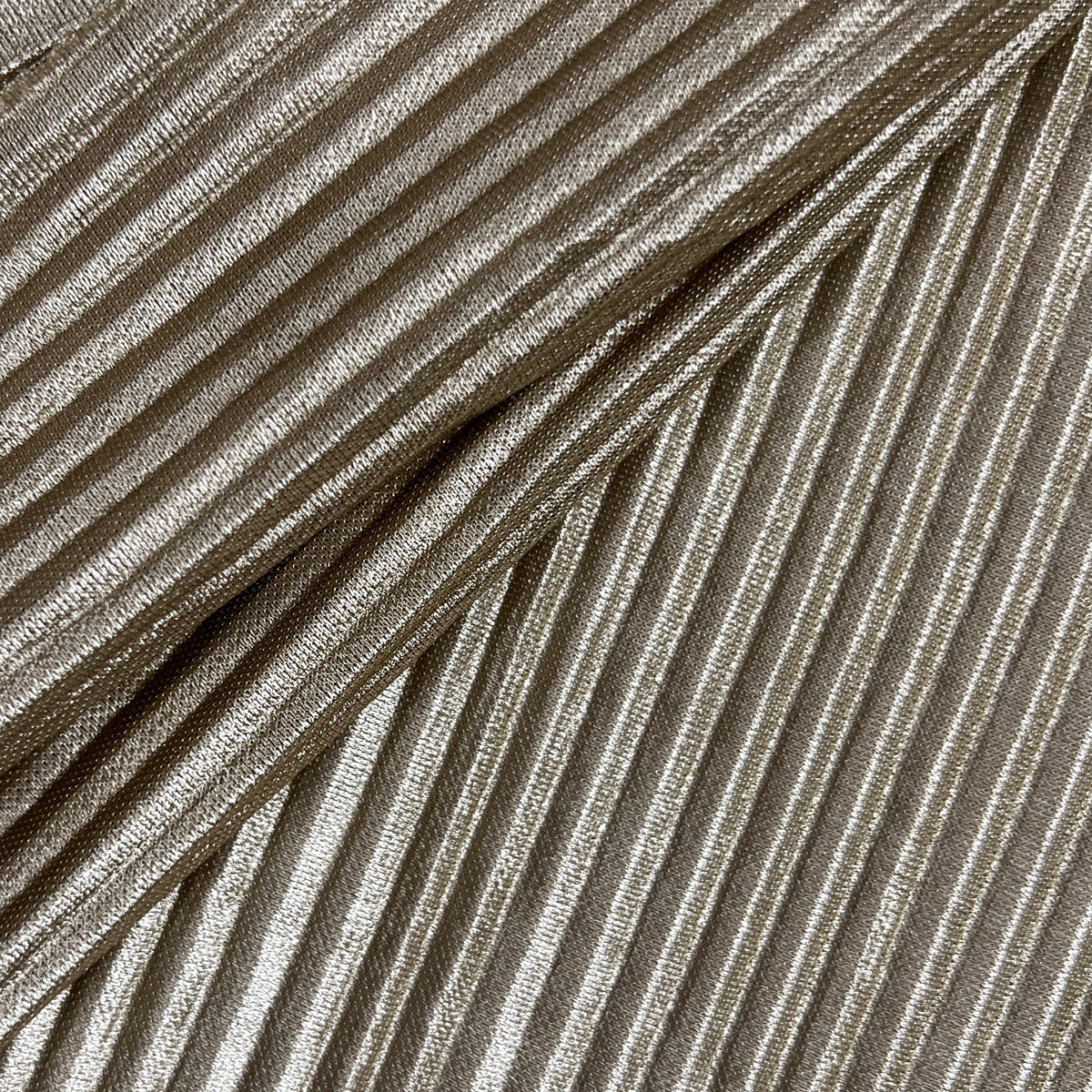 A close-up of a pleated fabric or material sample with narrow vertical pleats in a warm taupe-beige tone, photographed against a light neutral background.