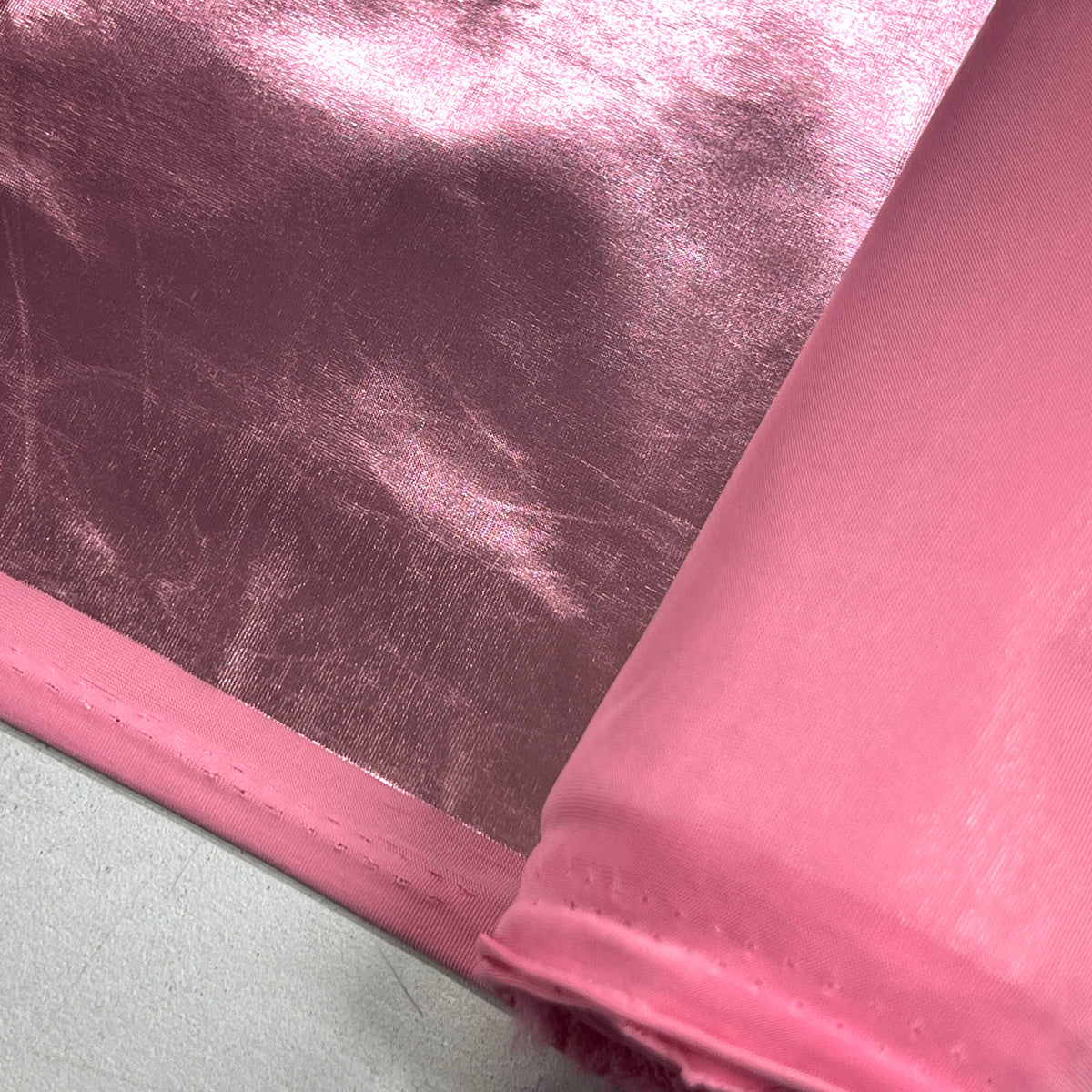 A close-up of shiny pink fabric, with one corner rolled back to show the material texture and edge, laid over a light gray surface.