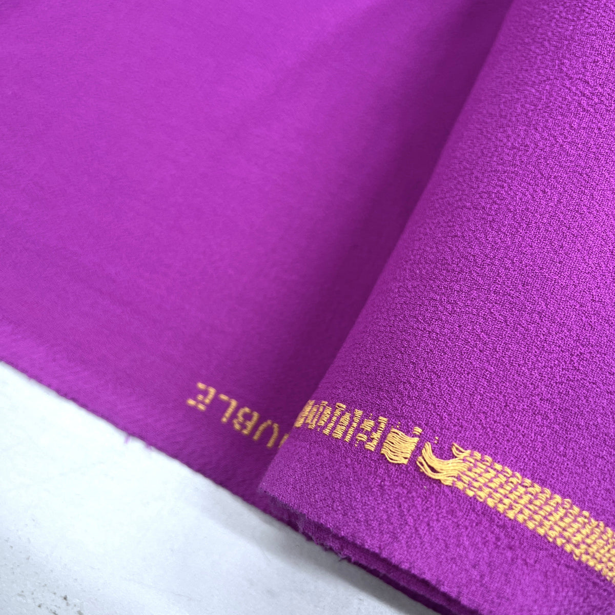 A close-up of a vibrant purple fabric, likely a saree, folded over to show its texture and border. The border features a gold woven stripe with a stylized emblem and partial text reading 'SAROO'.