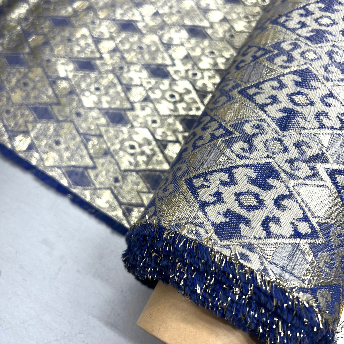 A close-up of a rolled piece of metallic fabric with a blue and silver geometric woven pattern and a fringed edge, mounted on a cardboard spool.