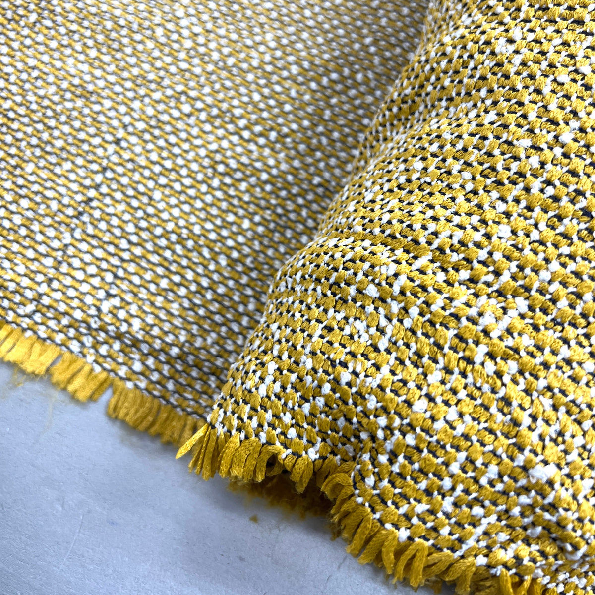 A close-up of a woven fabric with a textured basketweave pattern in mustard yellow and white, finished with a frayed edge.